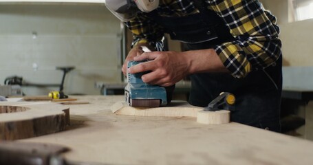 a man of European appearance in protective headphones and a mask processes a wood using a tool, sanding wood with a flex, work in a carpentry workshop close-up
