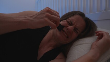 Woman Grimacing While Removing Black Mouth Tape While Lying in Bed in Dim Lighting, Discomfort Using Nighttime Tape Concept