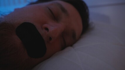Man Sleeping on Side with Black Mouth Tape in Dim Bedroom Light While Lying on White Pillow, Nose...