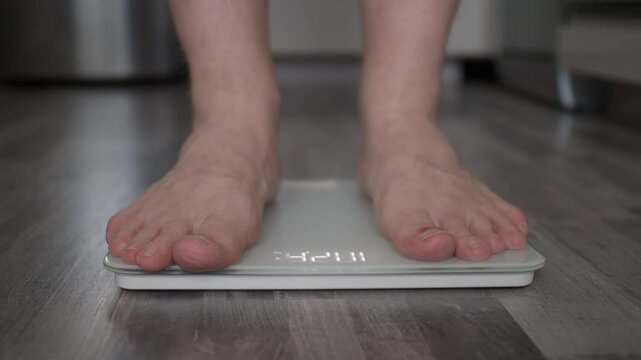 Men's feet step on scales to determine weight change, effectiveness of diet, exercise close up. Approach from afar, out of focus. Home environment, self-care health, wellness. Calm daytime gray shades
