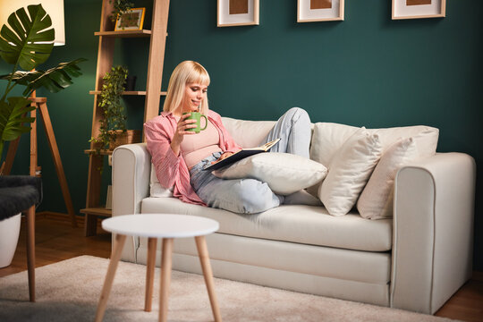 Serene young woman enjoys a quiet moment at home reading a book and sipping coffee on her comfortable sofa