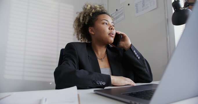 beautiful black girl with African hairstyle in a black business jacket, with an earring in the nose and a chain around her neck, seriously talking on the phone and typing in a laptop in a light office