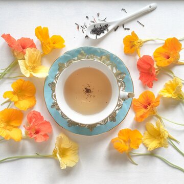 Overhead view of a Cup of Tea surrounded by yellow and orange nasturtium flowers with a teaspoon filled with loose tea leaves