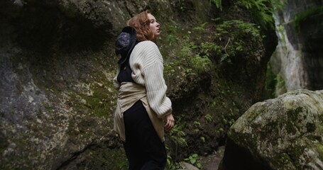 Russia, Caucasus. A young beautiful woman of European appearance, dressed in a warm sweater, makes her way through the stones in the cleft between the rocks overgrown with greenery