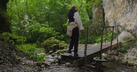 Fototapeta premium Russia, Caucasus. A young beautiful woman of European appearance crosses a fast mountain river on a log bridge. The beauty of the mountainous nature of the Caucasus