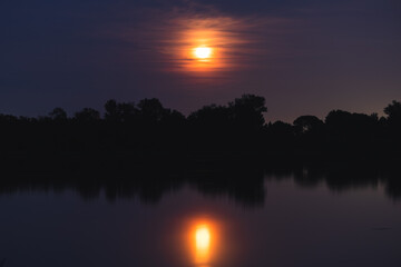 Obraz premium luna piena e di colore arancione che si specchia di notte sull'acqua tranquilla di una palude fluviale, in un ambiente naturale con alberi e cielo leggermente nuvoloso, in estate
