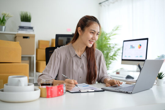Young woman analyzes sales performance and manages order details for her online business while seated in a modern workspace filled with packaging supplies and a laptop