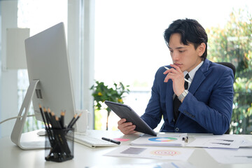 Young businessman holding a stylus and looking thoughtfully at a tablet surrounded by printed...