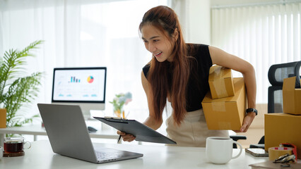 Confident young woman holding shipping boxes and checking order details on a laptop at her modern...