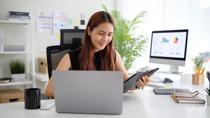 Smiling young businesswoman analyzing paperwork while working on laptop in a bright, modern office.