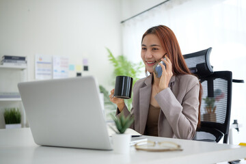 Fototapeta premium Young businesswoman holding coffee cup and talking on mobile phone at her modern office desk.