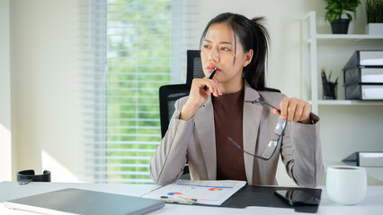 A professional woman in a modern office setting sits in deep thought, holding her eyeglasses in one hand and a pen in the other.