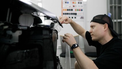 Professional mechanic meticulously applying protective vinyl wrap on vehicle headlights, carefully...