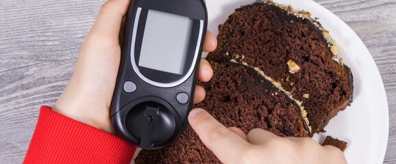 Hand of child holding glucometer, sweet chocolate cake. Checking sugar level during diabetes
