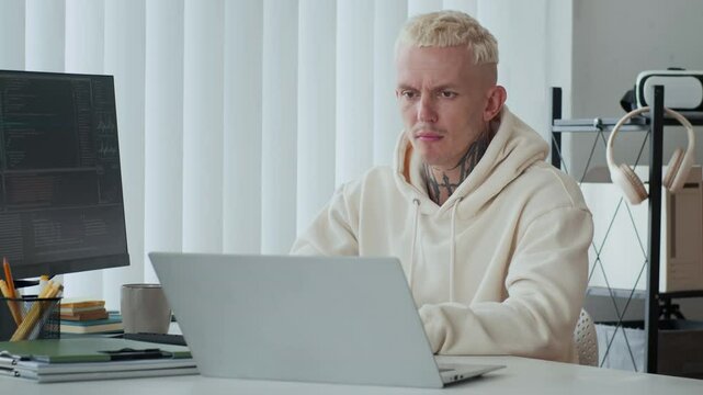 Medium shot of young Caucasian male tattooed developer analyzing coding screens on wireless laptop and computer monitor at office desk