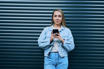 Stylish young woman in denim jacket looking down at smartphone while standing against dark...