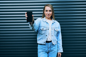 Young woman in denim jacket holding smartphone outwards with one hand and looking confidently at...