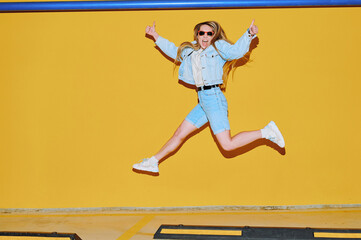 Energetic woman in denim outfit jumps mid-air showing thumbs up, excitement radiates from confident...