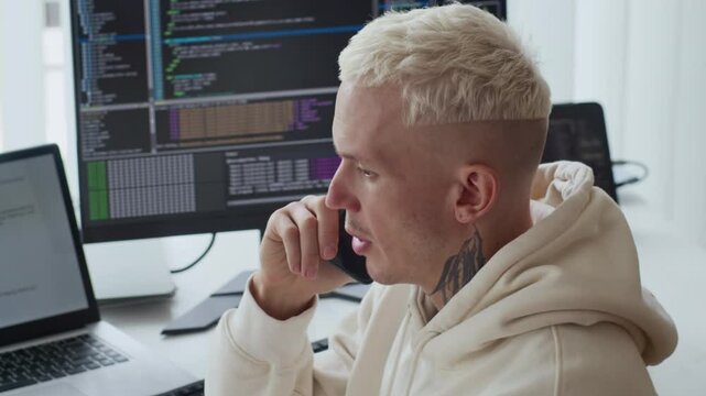 Side view of young male tattooed programmer talking on mobile phone while working on application update at office desk
