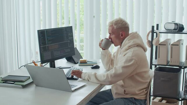 Side view of young male coder drinking tea while developing application for computers and laptops, sitting at office desk