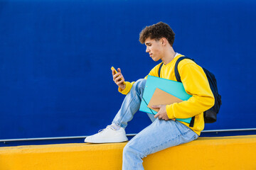 College student using smartphone while sitting on yellow wall with blue background