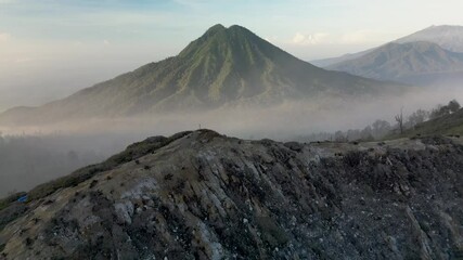 4K drone footage advancing along a crater edge trail at Ijen, Indonesia. Misty, cinematic atmosphere with people walking, crater walls, and a distant mountain in the fog. Captures nature, adventure, a - Powered by Adobe