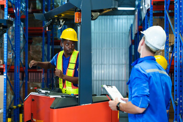 African male forklift operator wearing helmet and safety vest discussing logistics direction with white inspector holding clipboard during teamwork session inside warehouse area