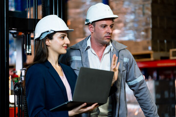 Male supervisor and female secretary wearing safety helmets having discussion while holding laptop inside warehouse during inspection and planning session with teamwork and communication