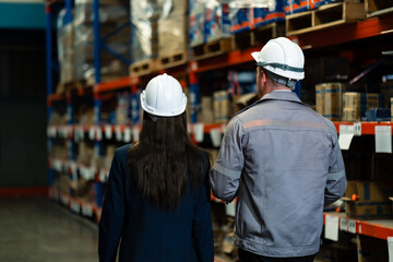 Warehouse manager and secretary walking side by side down storage aisle wearing helmets while monitoring product shelf area during warehouse inspection and internal stock control