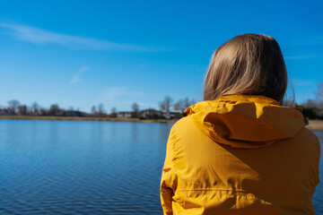 Fototapeta premium A young woman in a yellow jacket stands on the shore of a lake or river, gazing into the distance. Small town houses on the horizon. Peaceful countryside scenery. Sunny day
