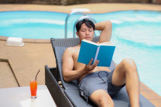 Young man lounging on a poolside chair, deeply immersed in a captivating book, with a refreshing cocktail close by, savoring a peaceful and relaxing day under the sun