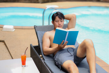 Young man enjoying his vacation, lying on a deck chair by the pool, reading a book and with a refreshing cocktail nearby, during a sunny day