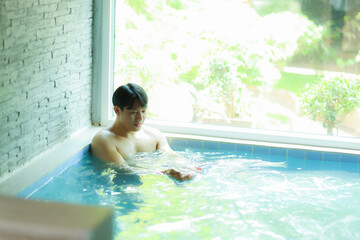 Asian tourist enjoying his summer vacation relaxing in a swimming pool inside a resort hotel, he is touching water with his hands and looking down