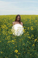 Fototapeta premium A nude woman stands in a blooming yellow flower field holding a round mirror, reflecting the flowers and sky in a surreal composition.