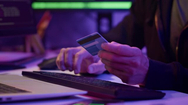 Close up shot of hands of anonymous hacker stealing users data holding credit card by laptop in neon room
