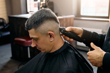 A man sits in a barbershop chair receiving a haircut. The barber uses clippers to create a fade...