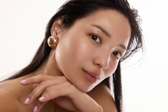Portrait of beautiful young Korean woman with smooth, radian, glowing skin looking directly into the camera, wearing golden massive earrings on white background. Skincare, cosmetics, jewelry concept