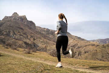Rear view of young unrecognizable woman trail running in the mountains on a partly cloudy day....
