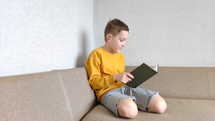 A boy with a hearing aid in his ear reads a book in his room