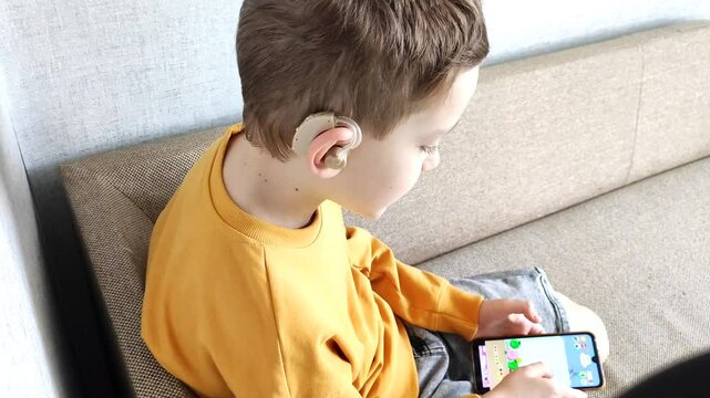 Boy with hearing aid in ear holds phone in hands, listens to audiobook while sitting in his room