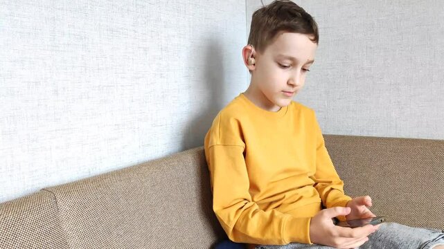 Boy with hearing aid in ear holds phone in hands, listens to audiobook while sitting in his room