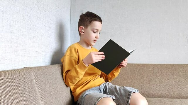 A boy with a hearing aid in his ear reads a book in his room