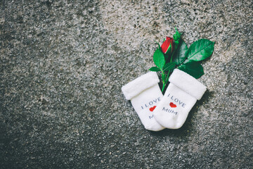 A pair of white baby socks with 'I LOVE MOM' text and a red heart lie beside a single red rose on a textured gray surface, symbolizing love and parenthood.