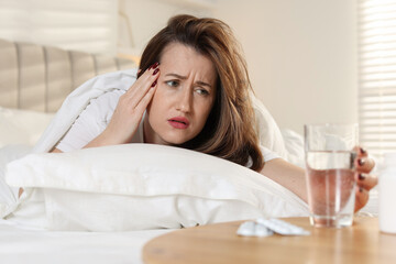 Unhappy woman with glass of water and pills suffering from hangover on bed at home