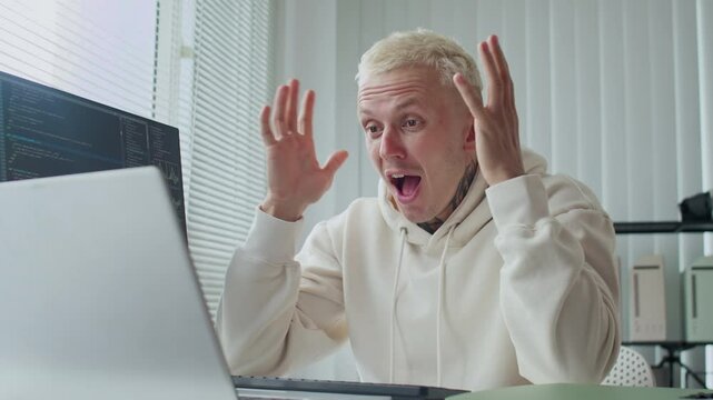 Medium shot of happy Caucasian tattooed man sitting in front of laptop screen while working with scripts at office desk