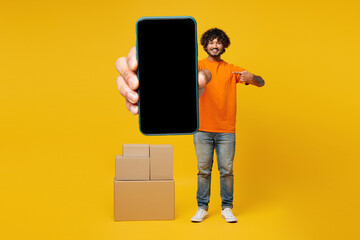 Full body young Indian man wear orange t-shirt casual clothes stand near stack cardboard boxes use...
