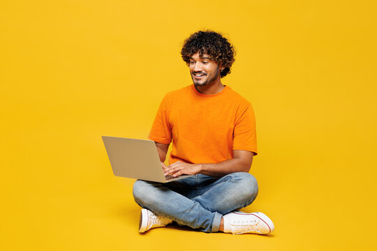 Full body young smart IT Indian man wear orange t-shirt casual clothes sit hold use work on laptop pc computer chatting online isolated on plain yellow background studio portrait. Lifestyle concept.