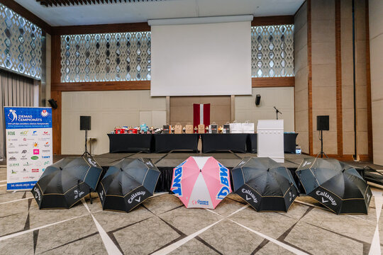 Indoor stage setup for an award event with branded umbrellas, sponsor board, and prizes arranged under a screen and Latvian flag backdrop..