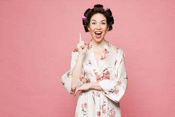 Young housewife housekeeper woman she wear white robe hair rollers holding index finger up with great new idea isolated on pastel light pink color background studio portrait. Morning routine concept.