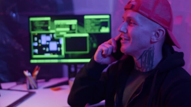 Medium shot of young Caucasian man in red cap with neck tattoo talking on mobile phone while sitting in neon-lit room with computer monitor displaying coding screen behind him
